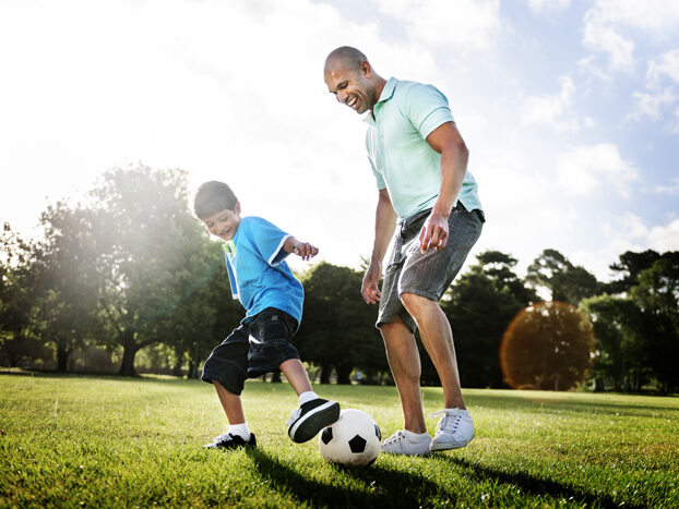 A father and son kick a soccer ball on green grass.